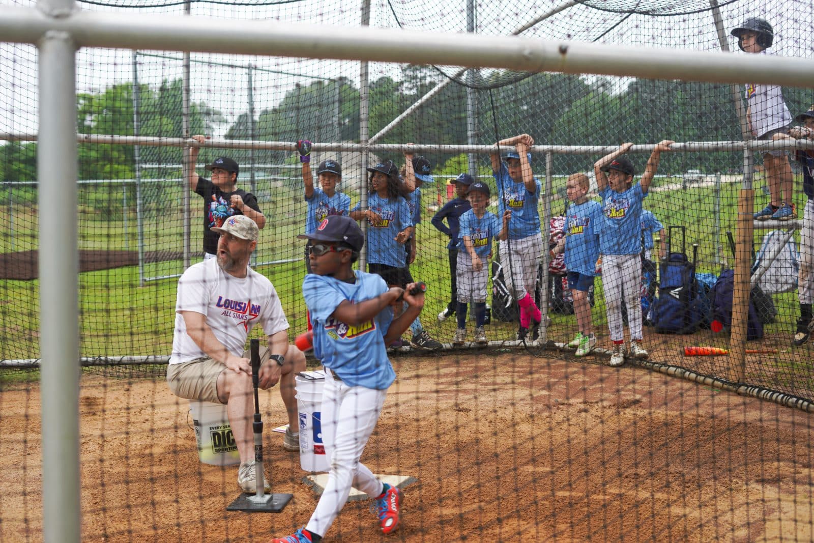 A coach working with young hitters in the batting cage as teammates watch.